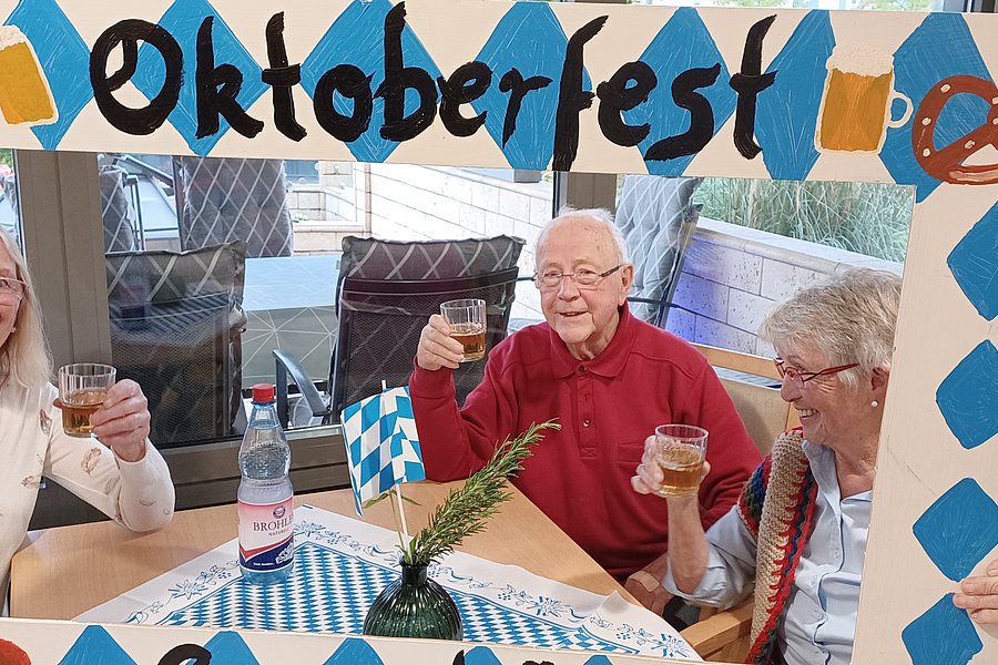 Vier Personen sitzen an einem Tisch mit blau-weiß kariertem Tischtuch und halten Biergläser, davor ein Rahmen mit der Aufschrift 'Oktoberfest' und Brezeln als Dekoration