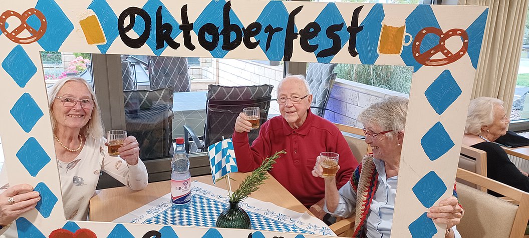 Vier Personen sitzen an einem Tisch mit blau-weiß kariertem Tischtuch und halten Biergläser, davor ein Rahmen mit der Aufschrift 'Oktoberfest' und Brezeln als Dekoration