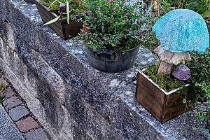 Blumentopf mit grünem Busch auf einer Steinmauer neben einem blauen Pilz aus Glas und weiteren Pflanzen