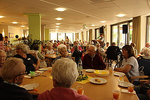 Mehrere ältere Menschen sitzen an Tischen in einem hellen Gemeinschaftsraum und hören einer stehenden Person zu