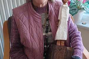 Ältere Frau mit grauen Locken, die glücklich sitzt und eine handgefertigte Puppe aus Holz hält.