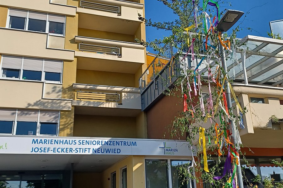 Mehrstöckiges Seniorenwohnheim mit Balkon und Eingangsbereich, davor ein geschmückter Maibaum mit bunten Bändern.