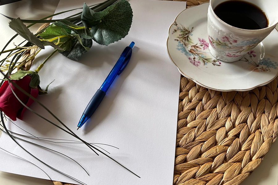 Auf einem geflochtenen Untersetzer liegt eine Tasse Kaffee auf einer Untertasse mit floralem Muster neben einem blauen Kugelschreiber und einer roten Rose auf weißem Papier