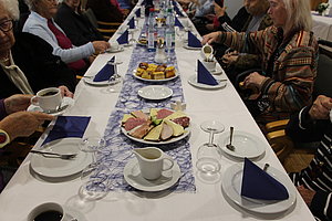 Langer gedeckter Tisch mit Tellern, Tassen, Gläsern, Servietten und Platten mit Aufschnitt und Kuchen, umgeben von sitzenden Personen
