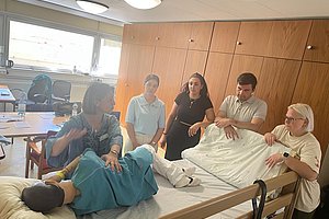 Schulungsszenario in einem Klassenzimmer mit fünf Personen um ein Bett mit einer simulierten Patientin.