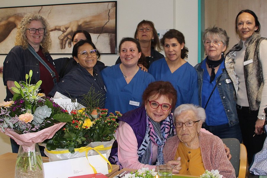 Gruppe von Menschen in einem Raum mit einem Tisch, auf dem Blumensträuße, Karten und ein kleiner Kuchen mit Kerze stehen.