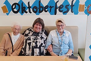 Drei sitzende Personen vor einer Wand mit blau-weißer Rauten-Dekoration, Brezeln, Biergläsern und dem Schriftzug 'Oktoberfest'