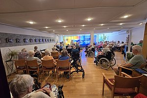 Sitzende ältere Menschen, viele im Rollstuhl, in einem großen Raum mit Holzboden und Deckenbeleuchtung, vorne ein bunter Wandbehang