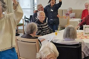 Aktive Senioren bei einer Veranstaltung, einige heben die Hände zur Teilnahme, fröhliche Umgebung.