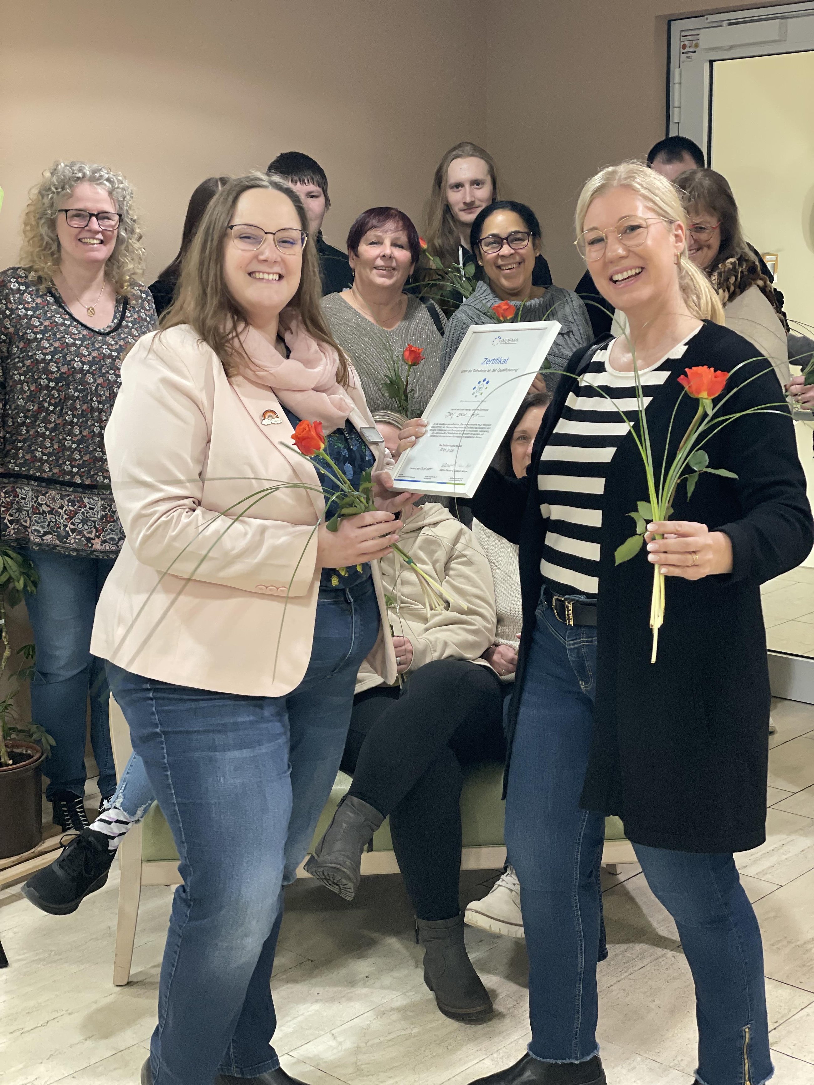 Zwei Frauen stehen, eine hält eine Urkunde, beide halten eine rote Rose, im Hintergrund mehrere Personen in einem Raum