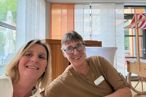 Zwei Personen sitzen an einem Tisch in einem hellen Raum mit großen Fenstern und einer Holzverkleidung im Hintergrund