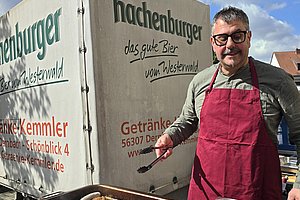 Mann mit roter Schürze steht vor zwei großen Tabletts mit gegrilltem Fleisch neben einem Getränkelieferwagen mit der Aufschrift 'Hachenburger das gute Bier vom Westerwald'