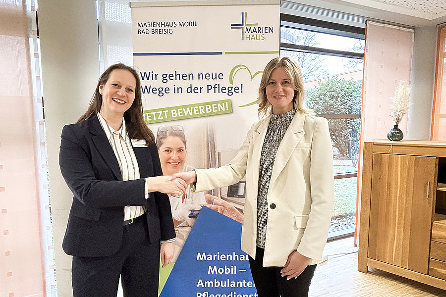 Zwei Frauen in Büro mit Fenster und Holzschrank schütteln sich die Hand vor einem Werbebanner für Marienhaus Mobil Ambulanten Pflegedienst