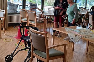 Innenraum mit Holzfußboden, mehreren Stühlen, Rollator und zwei Personen, die sich unterhalten