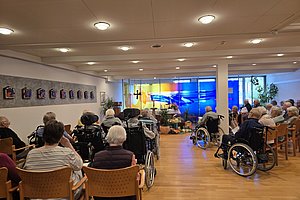 Senioren in Rollstühlen und Stühlen sitzen in einem Raum mit Holzboden und Deckenlampen und blicken auf eine bunte Wandprojektion