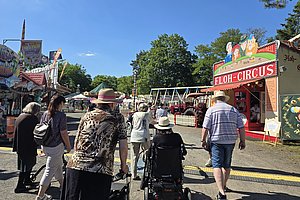 Menschen unterschiedlichen Alters gehen bei sonnigem Wetter auf einem Jahrmarktweg an einem Floh-Circus-Stand vorbei