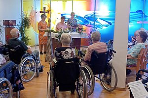 Mehrere ältere Menschen in Rollstühlen sitzen in einem Raum und hören zwei Personen zu, die vor einem farbigen Wandbild stehen