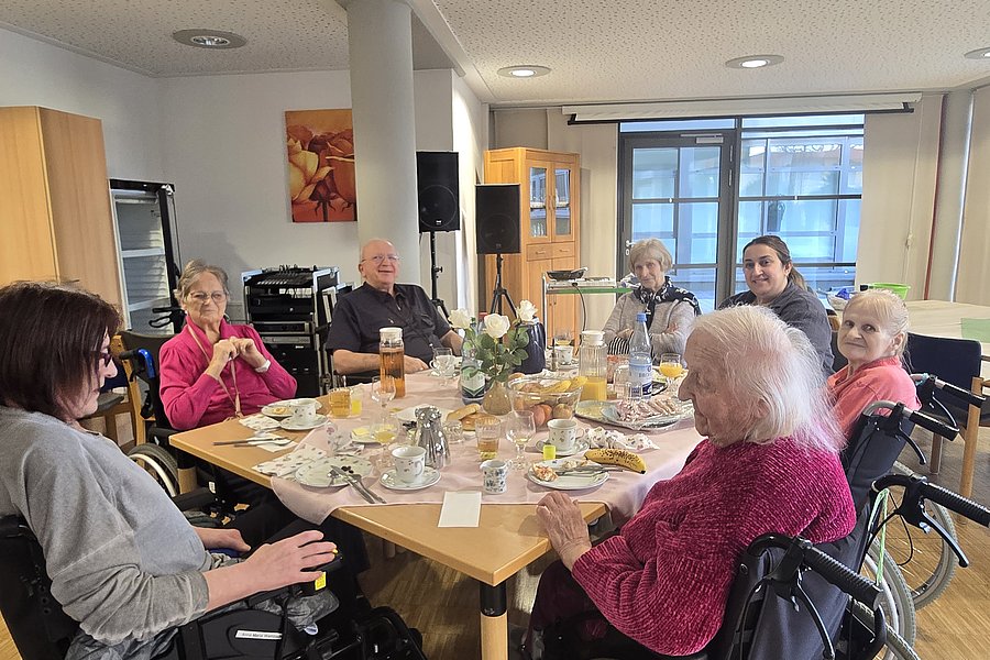 Geselliges Treffen im Aufenthaltsraum mit älteren Menschen, die in Rollstühlen am Tisch sitzen und Getränke genießen.
