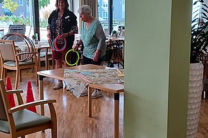 Zwei Personen stehen in einem Raum mit Holzboden und mehreren Stühlen, eine Person hält einen Ball