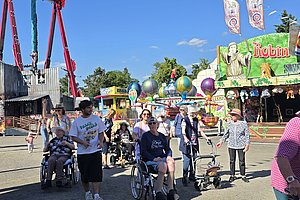 Besucher mit Rollstühlen auf einem bunten Jahrmarkt mit Fahrgeschäften und Festständen.