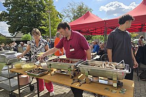 Drei Personen bedienen sich an einem Buffet mit Würstchen und Salat unter roten Zelten im Freien an einem sonnigen Tag