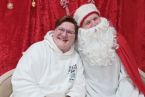 Zwei Personen sitzen vor einem roten Vorhang mit Weihnachtsdekoration, eine trägt einen weißen Kapuzenpullover, die andere ein Weihnachtsmannkostüm mit rotem Mantel und weißem Bart