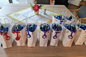 Mehrere weiße Papiertüten mit bunten Schlüsselanhängern und blauen Flaschen stehen auf einem Holztisch in einem Raum mit Stühlen und einem Tischläufer mit roter Blume