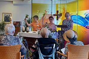 Mehrere ältere Menschen sitzen in einem Raum mit Stühlen und Tischen, im Hintergrund stehen zwei Personen vor einer farbigen Glaswand und sprechen.