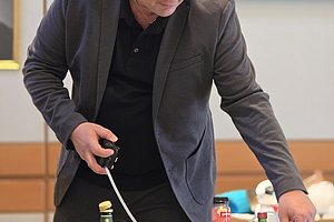 Person im Anzug mixt Getränke vor mehreren Flaschen und Bechern auf einem Tisch.
