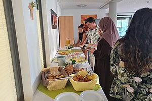 Mehrere Personen stehen an einem langen Tisch mit gedecktem Buffet in einem hellen Raum mit Fenster und Kreuz an der Wand
