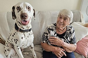 Ältere Frau mit Perlenkette und gemustertem Oberteil sitzt auf einem Sofa neben einem Dalmatiner mit offenem Mund
