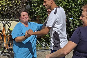 Drei Personen tanzen lachend im Freien auf gepflastertem Boden vor einem Gebäude mit Büschen im Hintergrund