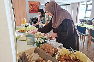 Buffet mit verschiedenen Speisen und Brötchen, Gäste servieren sich an einem gedeckten Tisch.
