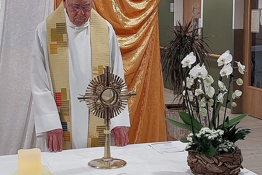 Ein Priester in weißem Gewand und Stola steht hinter einem mit weißem Tuch bedeckten Altar mit einem goldenen Monstranz-Objekt und einem Blumenarrangement aus weißen Orchideen