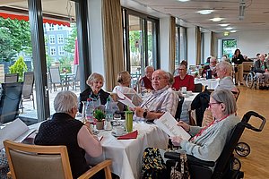 Mehrere ältere Menschen sitzen an Tischen in einem hellen Gemeinschaftsraum und halten Blätter in den Händen