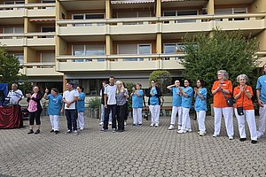 Gruppe von Menschen steht im Halbkreis vor einem mehrstöckigen Wohngebäude und klatscht