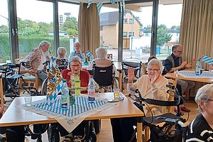 Mehrere ältere Menschen sitzen an Tischen in einem Gemeinschaftsraum mit großen Fenstern und Vorhängen, einige nutzen Rollatoren oder Rollstühle.