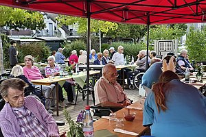 Mehrere ältere Menschen sitzen unter einem roten Pavillon an Tischen im Freien und unterhalten sich