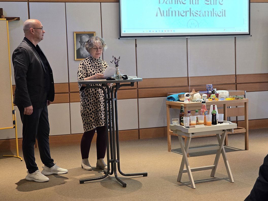 Zwei Personen bei einer Präsentation, Stehtisch mit verschiedenen Flaschen und Utensilien im Vordergrund.