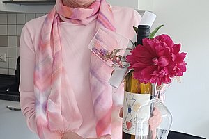 Frau mit rosa Schal und Pullover hält eine Weinflasche mit roter Blume und zwei Weingläsern in der Hand