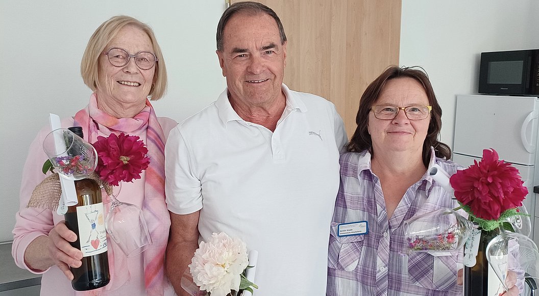 Drei Personen stehen nebeneinander, zwei Frauen halten rote Blumen und eine Flasche, der Mann in der Mitte hält eine weiße Blume