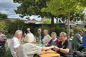 Gruppe älterer Menschen sitzt an einem Holztisch im Freien unter Bäumen und trinkt gemeinsam