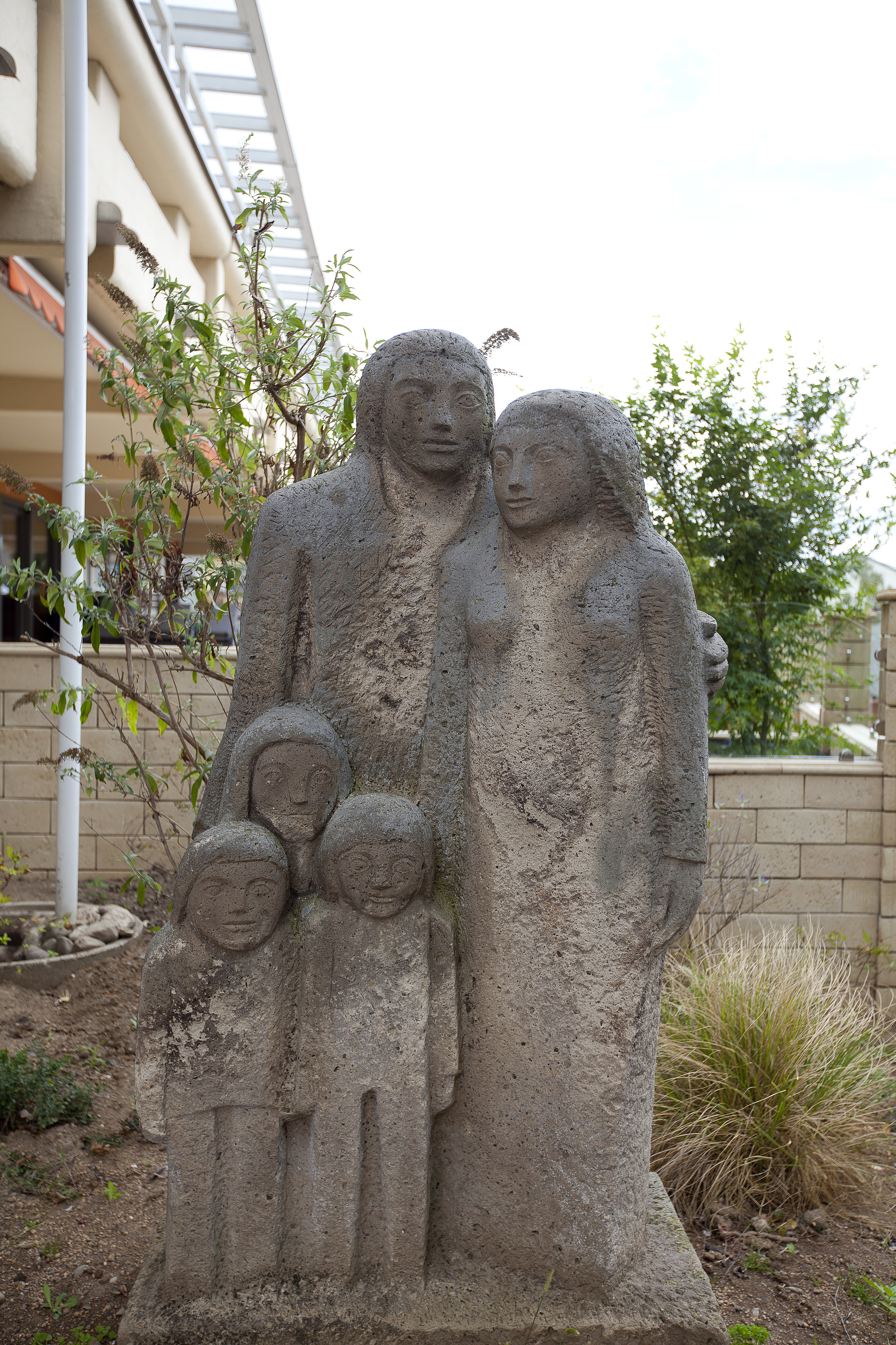 Steinerne Skulptur einer stehenden Familie mit zwei Erwachsenen und zwei Kindern in einem Garten mit Bäumen und Haus im Hintergrund.
