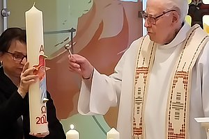 Priester in weißem Gewand mit besticktem Stola hält ein liturgisches Werkzeug vor eine große weiße Kerze mit roten Symbolen und Jahreszahl