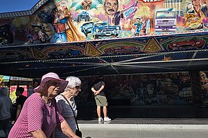 Zwei Frauen mit Rollator vor einem bunten Schaustellerstand, umgeben von nostalgischen Autos und lebhaften Bildern.