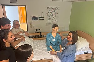 Fünf Personen sitzen und stehen um ein Krankenbett, in dem eine medizinische Übungspuppe sitzt, an der eine Frau mit Brille und blauer Bluse eine Übung durchführt.