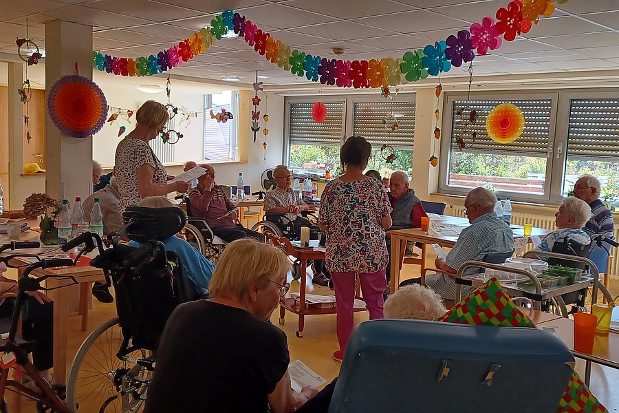 Mehrere ältere Menschen sitzen in einem hellen Raum mit großen Fenstern, einige im Rollstuhl, während zwei Frauen stehen und sich mit ihnen unterhalten, bunte Girlanden und Dekorationen hängen von der Decke