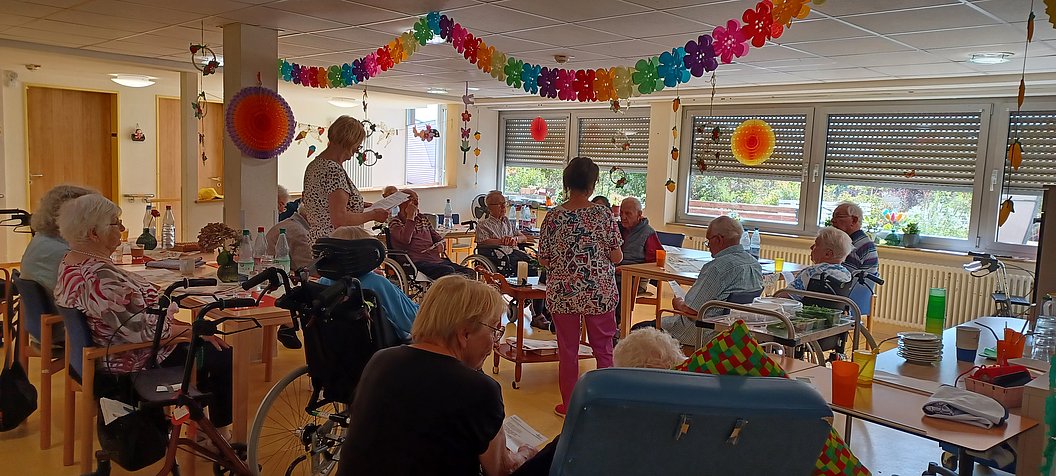 Mehrere ältere Menschen sitzen in einem hellen Raum mit großen Fenstern, einige im Rollstuhl, während zwei Frauen stehen und sich mit ihnen unterhalten, bunte Girlanden und Dekorationen hängen von der Decke