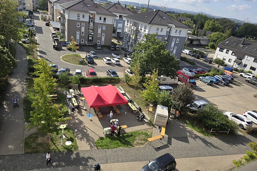 Luftaufnahme eines Wohngebiets mit Parkplätzen, Bäumen und einem roten Pavillon auf einem Platz