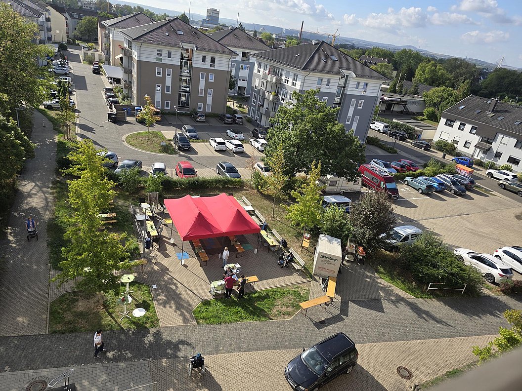 Luftaufnahme eines Wohngebiets mit Parkplätzen, Bäumen und einem roten Pavillon auf einem Platz